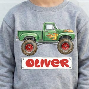 May include: A gray sweatshirt featuring a green monster truck graphic with flames and the name "OLIVER" in red. The truck has large tires with red rims. The sweatshirt is made of a soft material.