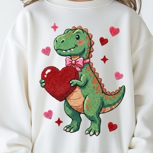 May include: A white sweatshirt featuring an embroidered green dinosaur holding a red heart. The dinosaur has an orange spine and a pink bow tie. Pink hearts and stars are scattered around the dinosaur, creating a Valentine's Day theme.