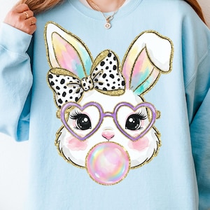 May include: Light blue sweatshirt featuring a cartoon bunny face. The bunny has heart-shaped glasses, a polka-dot bow, and is blowing a bubble. The bunny's ears have a rainbow gradient and gold glitter trim.