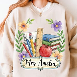 May include: Beige sweatshirt featuring a watercolor design for a teacher. The design includes books, an apple, pencils, a ruler, and the text "Mrs. Amelia" in a decorative frame, surrounded by floral elements.