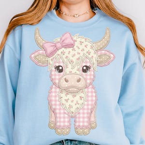 May include: A light blue sweatshirt featuring a cartoon illustration of a Highland cow. The cow has a pink bow, floral accents, and a pink and white gingham pattern. The cow's horns and ears are tan.