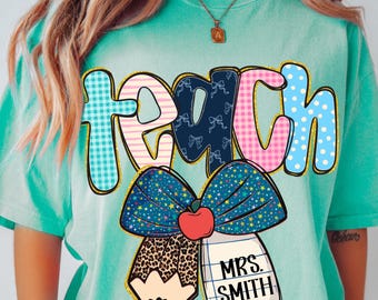 May include: A turquoise t-shirt featuring the word "TEACH" in a vibrant, playful font. The letters are decorated with patterns including gingham, polka dots, and bows. A large bow graphic with a pencil and apple is below the word, with the name "MRS. SMITH" on a notepad.