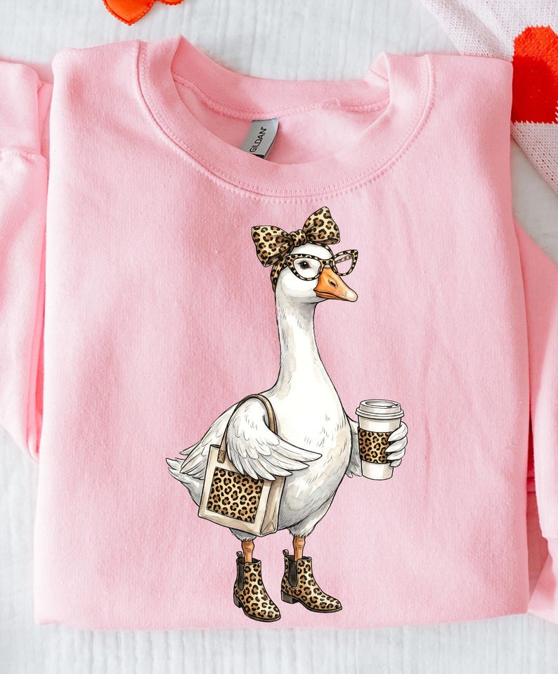 May include: Pink sweatshirt featuring a whimsical illustration of a white goose wearing glasses, a leopard print bow, and boots. The goose carries a tote bag and a coffee cup, both with leopard print accents.