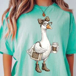 May include: A teal t-shirt featuring a whimsical illustration of a goose wearing glasses, a bow, and boots, holding a coffee cup and a tote bag, all with a leopard print design. The goose is the central focus of the graphic.