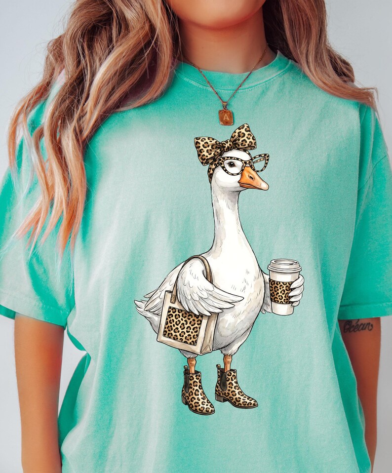 May include: A teal t-shirt featuring a whimsical illustration of a goose wearing glasses, a bow, and boots, holding a coffee cup and a tote bag, all with a leopard print design. The goose is the central focus of the graphic.