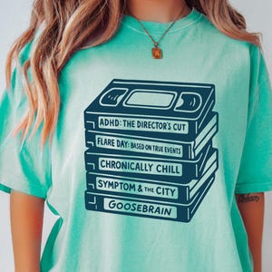 Op de afbeelding: Een licht-turquoise t-shirt met een marineblauwe afbeelding van gestapelde VHS-banden, elk met een humoristische titel zoals "ADHD: The Director's Cut" en "Goosebrain". Het shirt heeft een relaxte pasvorm en korte mouwen.