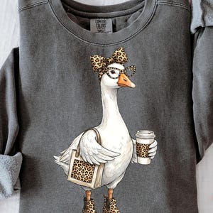 May include: Dark gray Comfort Colors sweatshirt featuring a whimsical illustration of a goose wearing glasses and a leopard print bow, boots, and carrying a tote bag and coffee cup with a leopard print design.