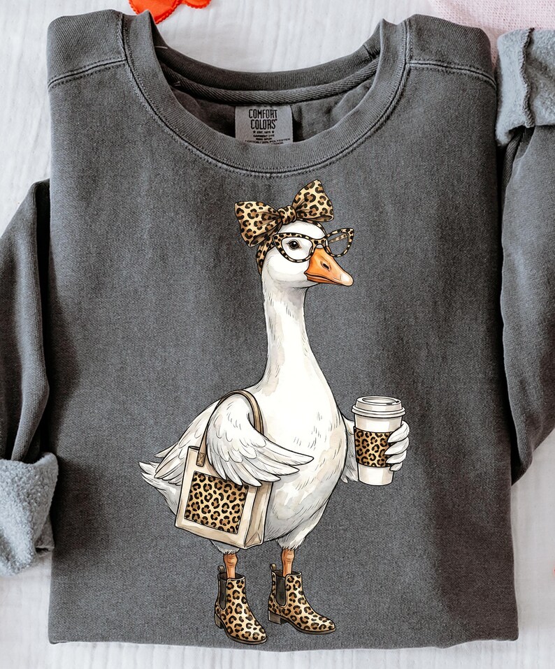 May include: Dark gray Comfort Colors sweatshirt featuring a whimsical illustration of a goose wearing glasses and a leopard print bow, boots, and carrying a tote bag and coffee cup with a leopard print design.