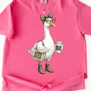 May include: A pink Comfort Colors t-shirt featuring a whimsical illustration of a white goose wearing glasses, a bow, and leopard print boots. The goose carries a tote bag and a coffee cup, both with leopard print accents.