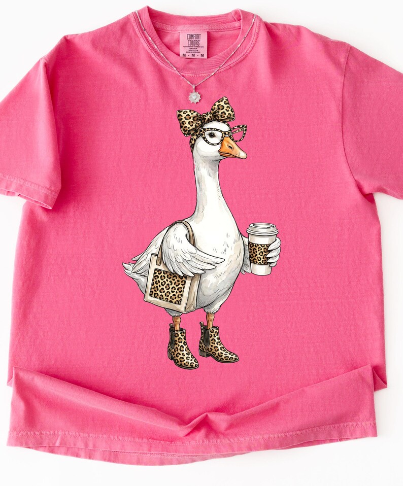 May include: A pink Comfort Colors t-shirt featuring a whimsical illustration of a white goose wearing glasses, a bow, and leopard print boots. The goose carries a tote bag and a coffee cup, both with leopard print accents.