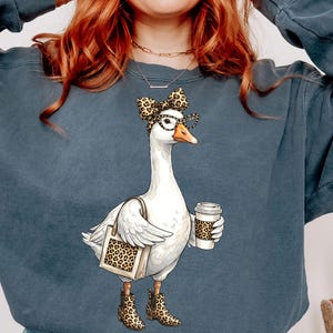 May include: A blue-grey sweatshirt featuring a whimsical illustration of a goose wearing glasses, a bow, and boots, all with a leopard print design. The goose is carrying a tote bag and a coffee cup, also with leopard print.