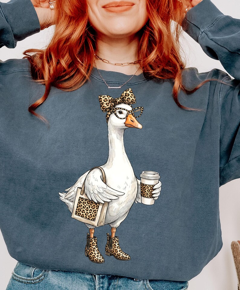 May include: A blue-grey sweatshirt featuring a whimsical illustration of a goose wearing glasses, a bow, and boots, all with a leopard print design. The goose is carrying a tote bag and a coffee cup, also with leopard print.