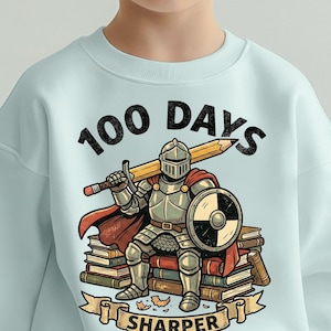 Puede incluir: Sudadera azul claro con un dibujo de un caballero con una espada-lápiz y un escudo, sentado sobre libros. El texto dice "100 DAYS SHARPER".