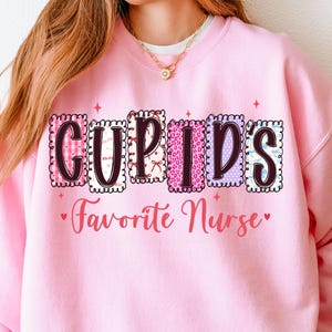 Puede incluir: Sudadera rosa con el texto "Cupid's Favorite Nurse" en una fuente decorativa. La palabra "Cupid's" está en un estilo de letras en bloque con varios patrones. El texto es de color rosa con un diseño de corazón.