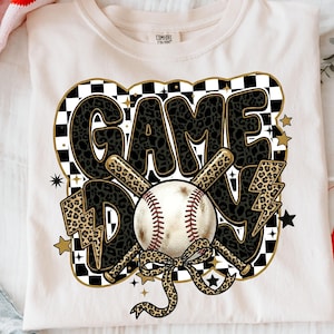 Può includere: T-shirt color crema con grafica a tema baseball. Il design presenta le parole "GAME DAY" in un carattere con stampa leopardata nera, mazze da baseball incrociate, una palla da baseball e fulmini. Stelle dorate e uno sfondo a scacchi completano il design.