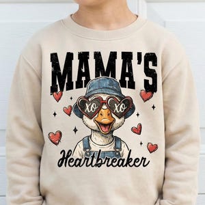 Puede incluir: Sudadera beige con un pato de dibujos animados con gafas de sol en forma de corazón y un sombrero de mezclilla. El texto "MAMA'S Heartbreaker" está impreso en la parte delantera, rodeado de corazones rojos.