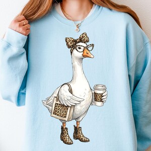 May include: Light blue sweatshirt featuring a cartoon goose wearing glasses, a leopard print bow, and boots. The goose is holding a coffee cup and a tote bag with a leopard print design. The goose is the central focus of the design.