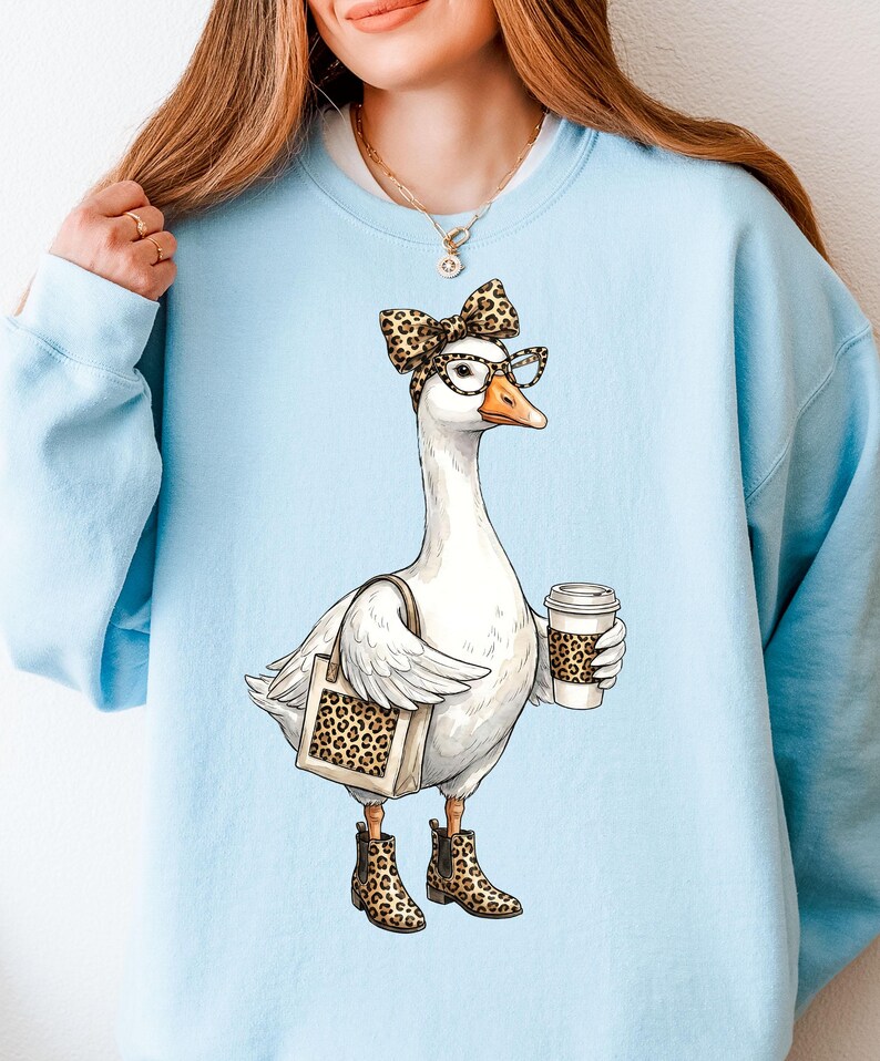 May include: Light blue sweatshirt featuring a cartoon goose wearing glasses, a leopard print bow, and boots. The goose is holding a coffee cup and a tote bag with a leopard print design. The goose is the central focus of the design.