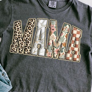 May include: Dark gray t-shirt with the word "MAMA" in a collegiate style. The letters are filled with various patterns: leopard print, marble, floral, and a brown and white checkerboard design. The shirt has a crew neck and a Comfort Colors tag.