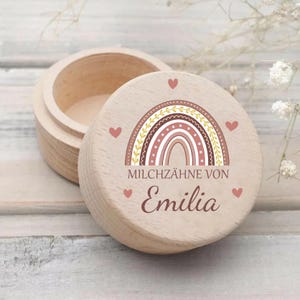 Könnte beinhalten: Eine kleine, offene Holzkiste mit Deckel, verziert mit einem Regenbogen-Design in Braun, Rosa und Gelb. Der Deckel trägt auch den Text "MILCHZÄHNE VON Emilia" und kleine rosa Herzen. Die Kiste ist aus hellem Holz.