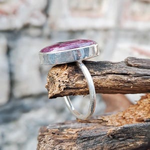 May include: A silver ring featuring a large oval pink gemstone. The ring is displayed on a piece of driftwood.