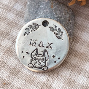 Könnte beinhalten: Ein silberner, runder Hundeanhänger mit dem eingravierten Namen "Max". Darunter befindet sich eine detaillierte Illustration einer Französischen Bulldogge. Der Anhänger ist mit Blattmotiven und kleinen sternförmigen Akzenten verziert.