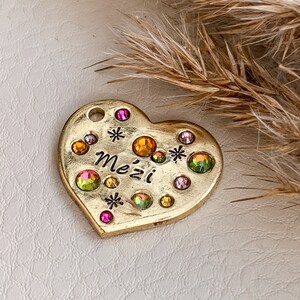 May include: A heart-shaped, gold-colored pendant with the word "Mezi" in black script. The pendant is adorned with colorful, iridescent gemstones and small star-shaped accents. The pendant has a small hole at the top.