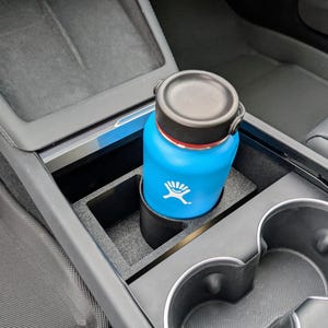 May include: A blue water bottle with a black lid and a white logo is in a car cup holder. The cup holder is part of a black car console.