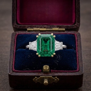 Puede incluir: Un anillo con una gran piedra preciosa verde de corte esmeralda rectangular engastada en garras de oro amarillo, flanqueada por dos diamantes transparentes de corte baguette a cada lado, sobre una banda plateada en un estuche de terciopelo.