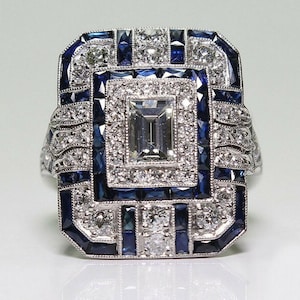 May include: An Art Deco-style ring with a rectangular face. The ring features a large, rectangular, clear center stone, surrounded by smaller white and blue stones. The setting is silver-toned, and the design is symmetrical and ornate.