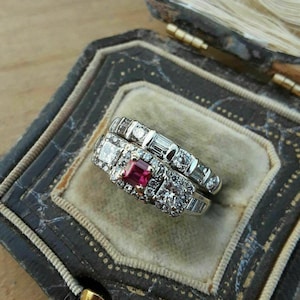 May include: A close-up of a ring set with a red gemstone surrounded by smaller diamonds. The ring is in a velvet-lined box.