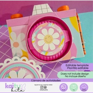 May include: A paper craft camera template featuring a pink body with a white daisy and smiley face design on the lens. The camera includes orange knobs, a grid pattern on top, and an orange crayon attached to the side. Text reads Editable template.