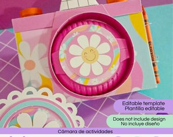 May include: A paper craft camera template featuring a pink body with a white daisy and smiley face design on the lens. The camera includes orange knobs, a grid pattern on top, and an orange crayon attached to the side. Text reads Editable template.