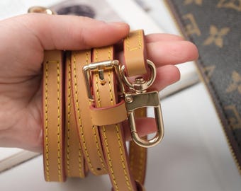 Pode incluir: Alça de bolsa de couro bege com ferragens douradas, incluindo uma fivela e um fecho giratório. A alça tem costuras amarelas e um interior vermelho. O fundo está desfocado.