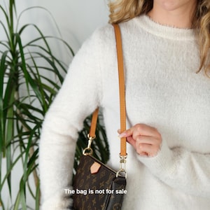 May include: Brown monogrammed shoulder bag with a tan leather strap and gold-colored hardware. The bag is held by a person wearing a white sweater. The text on the bag says "The bag is not for sale."