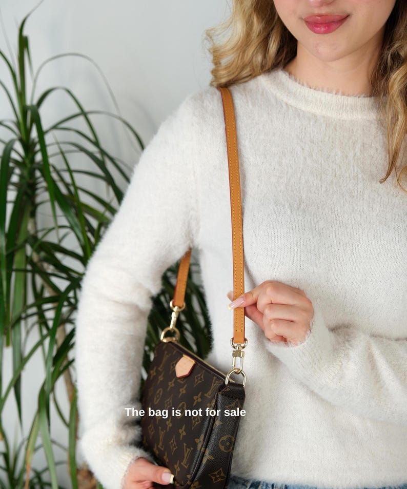 May include: Brown monogrammed shoulder bag with a tan leather strap and gold-colored hardware. The bag is held by a person wearing a white sweater. The text on the bag says "The bag is not for sale."