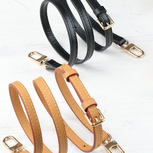 May include: Two leather purse straps are shown, one black and one tan. The black strap features gold-coloured hardware, including a buckle and a clasp. The tan strap has a similar design with gold-coloured hardware and a red interior.