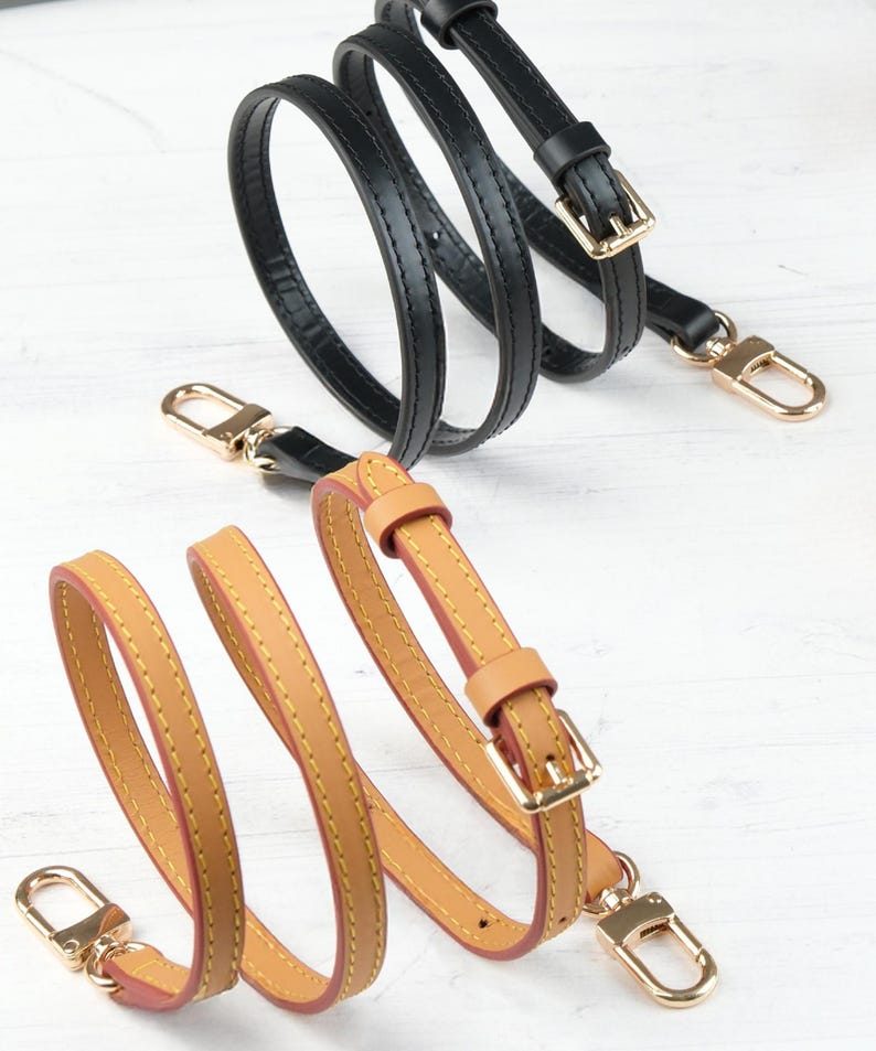 May include: Two leather purse straps are shown, one black and one tan. The black strap features gold-coloured hardware, including a buckle and a clasp. The tan strap has a similar design with gold-coloured hardware and a red interior.