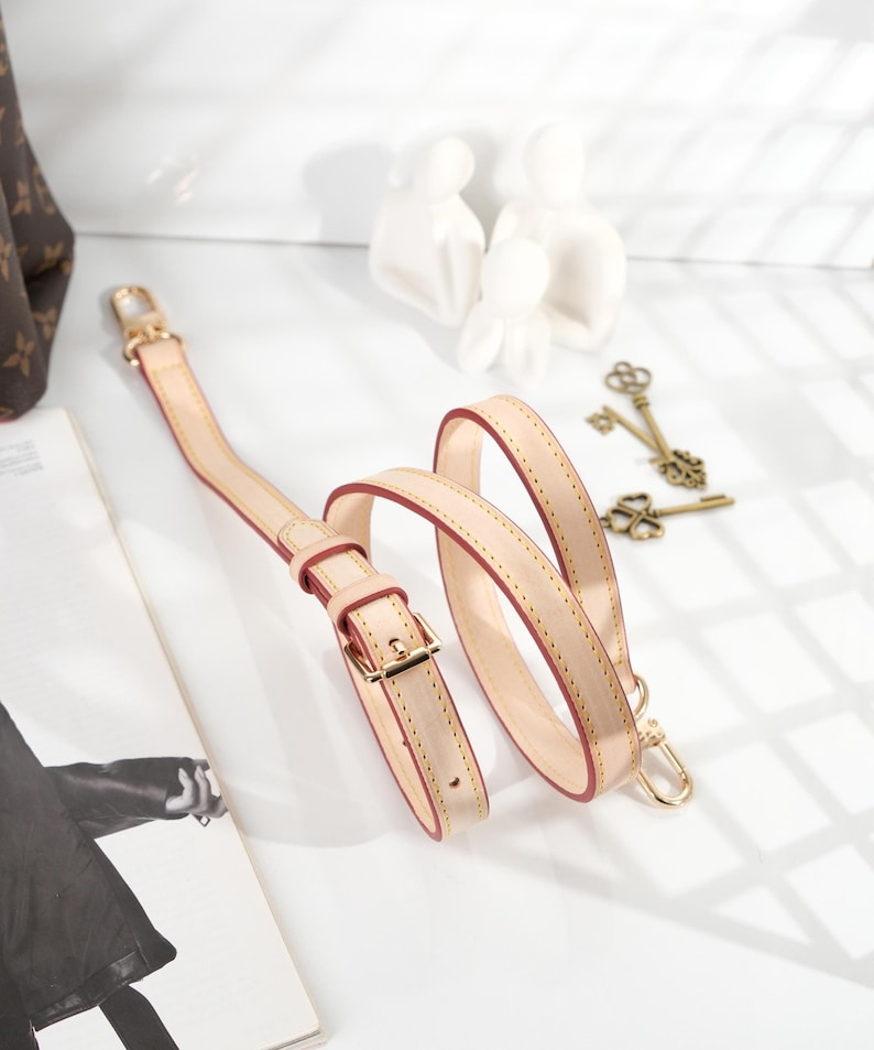 May include: A beige leather strap with a gold buckle and two gold clasps. The strap is folded and coiled on a white surface.