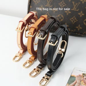 May include: Four leather purse straps in cream, tan, dark brown, and black. Each strap has a gold-coloured buckle and clasp. The bag in the background is not for sale.
