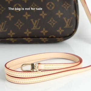 May include: A light beige leather purse strap with gold-colored hardware. The strap is thin and has a red edge. The background shows a brown purse with a repeating pattern and the text "The bag is not for sale".