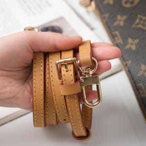 May include: Tan leather purse strap with gold-tone hardware, including a buckle and a swivel clasp. The strap is stitched with yellow thread and has a red edge. The strap is shown next to a brown purse with a monogram pattern.