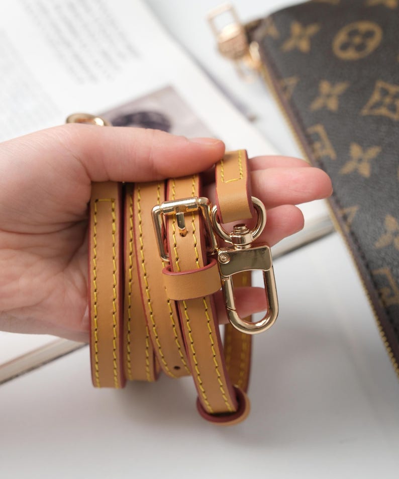 May include: Tan leather purse strap with gold-tone hardware, including a buckle and a swivel clasp. The strap is stitched with yellow thread and has a red edge. The strap is shown next to a brown purse with a monogram pattern.