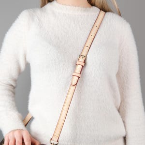 May include: A light brown leather shoulder strap with a gold buckle. The strap is attached to a white fuzzy sweater.