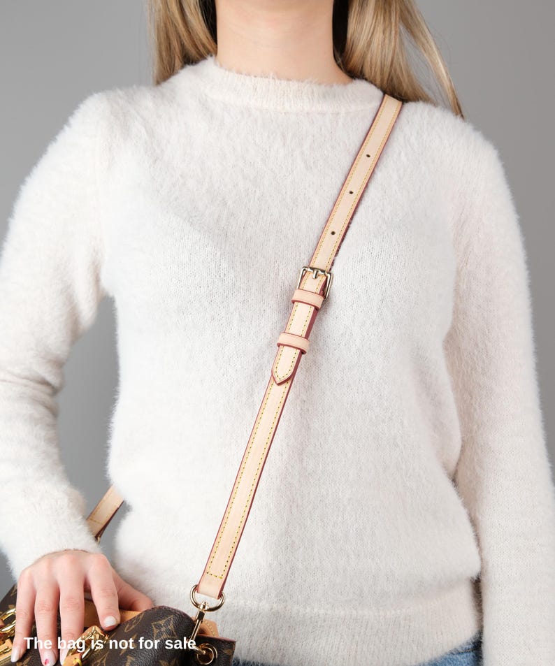 May include: A light brown leather shoulder strap with a gold buckle. The strap is attached to a white fuzzy sweater.