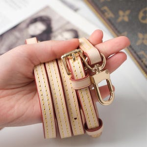 May include: A cream leather purse strap with gold-tone metal hardware. The strap has a buckle, a swivel clasp, and yellow stitching. The strap is folded and held in a hand.