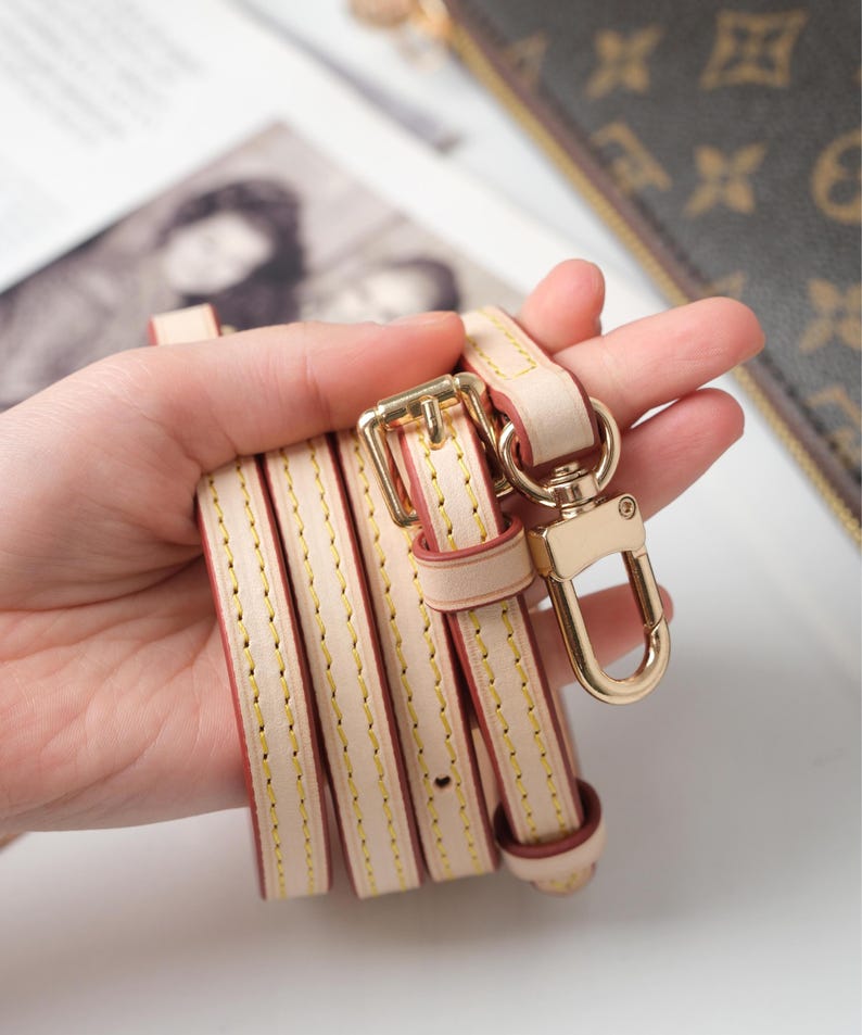 May include: A cream leather purse strap with gold-tone metal hardware. The strap has a buckle, a swivel clasp, and yellow stitching. The strap is folded and held in a hand.