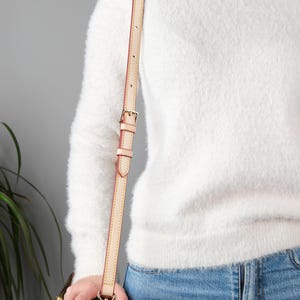 May include: A light brown leather shoulder strap with a gold buckle. The strap is attached to a brown leather bag.