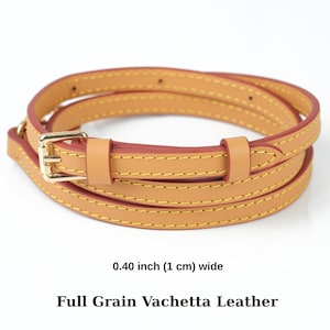 May include: A tan leather strap with a gold-coloured buckle. The strap is 1 cm wide and has yellow stitching. The text "Full Grain Vachetta Leather" is below the strap.