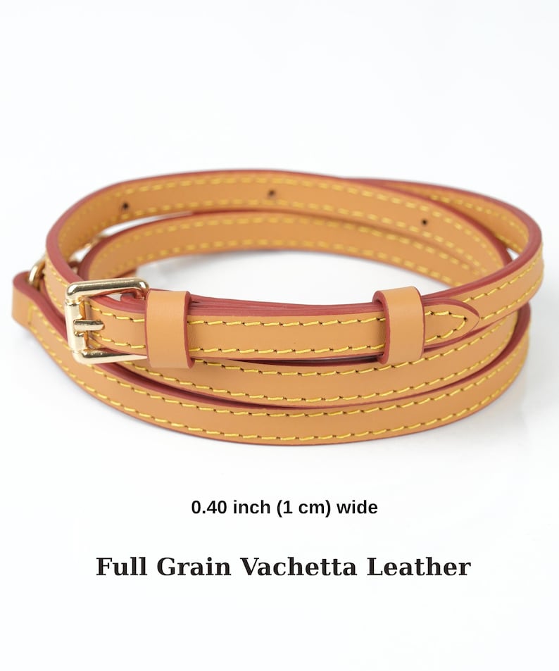 May include: A tan leather strap with a gold-coloured buckle. The strap is 1 cm wide and has yellow stitching. The text "Full Grain Vachetta Leather" is below the strap.